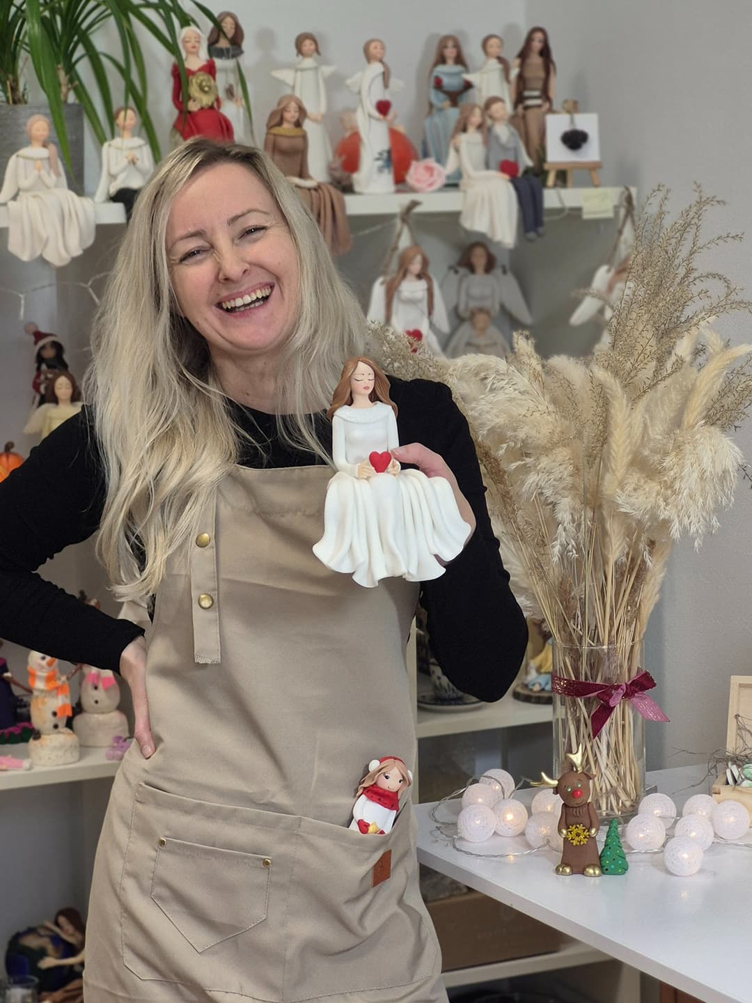 Radosna Marta Sowa w beżowym fartuchu prezentuje figurkę z zimnej porcelany przedstawiającą postać anioła trzymającego czerwone serce. W tle widać półki pracowni wypełnione dziesiątkami podobnych, ręcznie robionych figurek aniołów, zachęcających do wzięcia udziału w jednodniowym warsztacie dla kobiet podstawy zimnej porcelany w Ośrodku Rozwoju dla Kobiet Żeglarska Przystań.