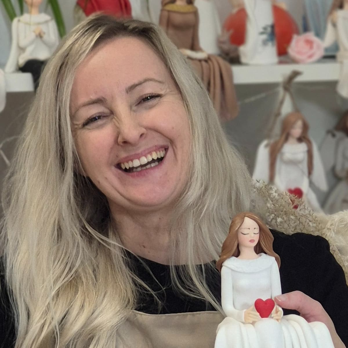 Uśmiechnięta Marta Sowa, prowadząca jednodniowe warsztaty dla kobiet z podstaw zimnej porcelany, w fartuchu rzemieślniczym. Trzyma w dłoniach gotową figurkę z zimnej porcelany – kobietę w białej sukni z czerwonym sercem. W tle widać półki pełne innych, ręcznie robionych aniołków i postaci.
